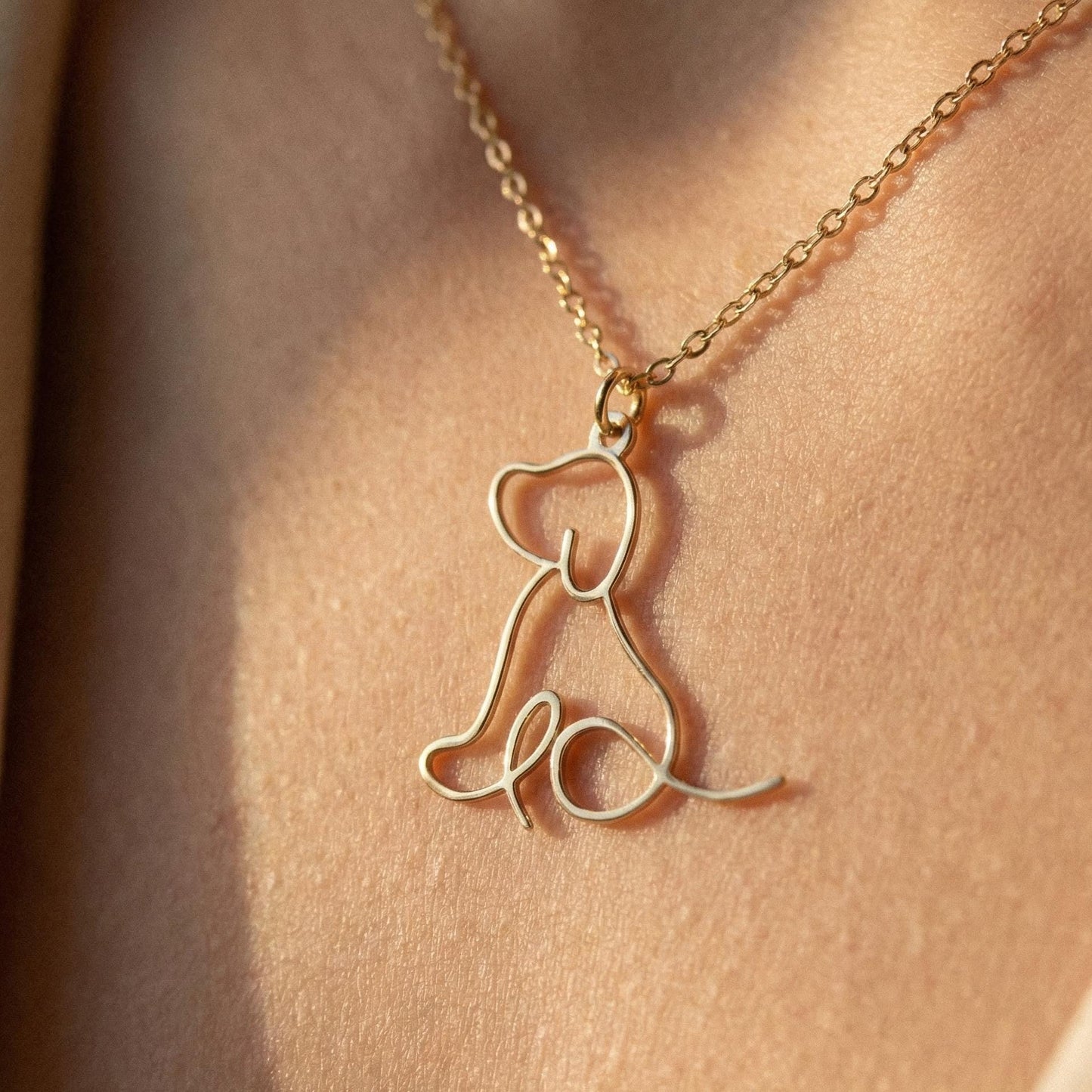 May include: A gold-colored necklace featuring a pendant in the shape of a sitting dog. The dog is depicted in a minimalist, line-art style. The necklace is worn against a skin-toned background, highlighting the delicate design.