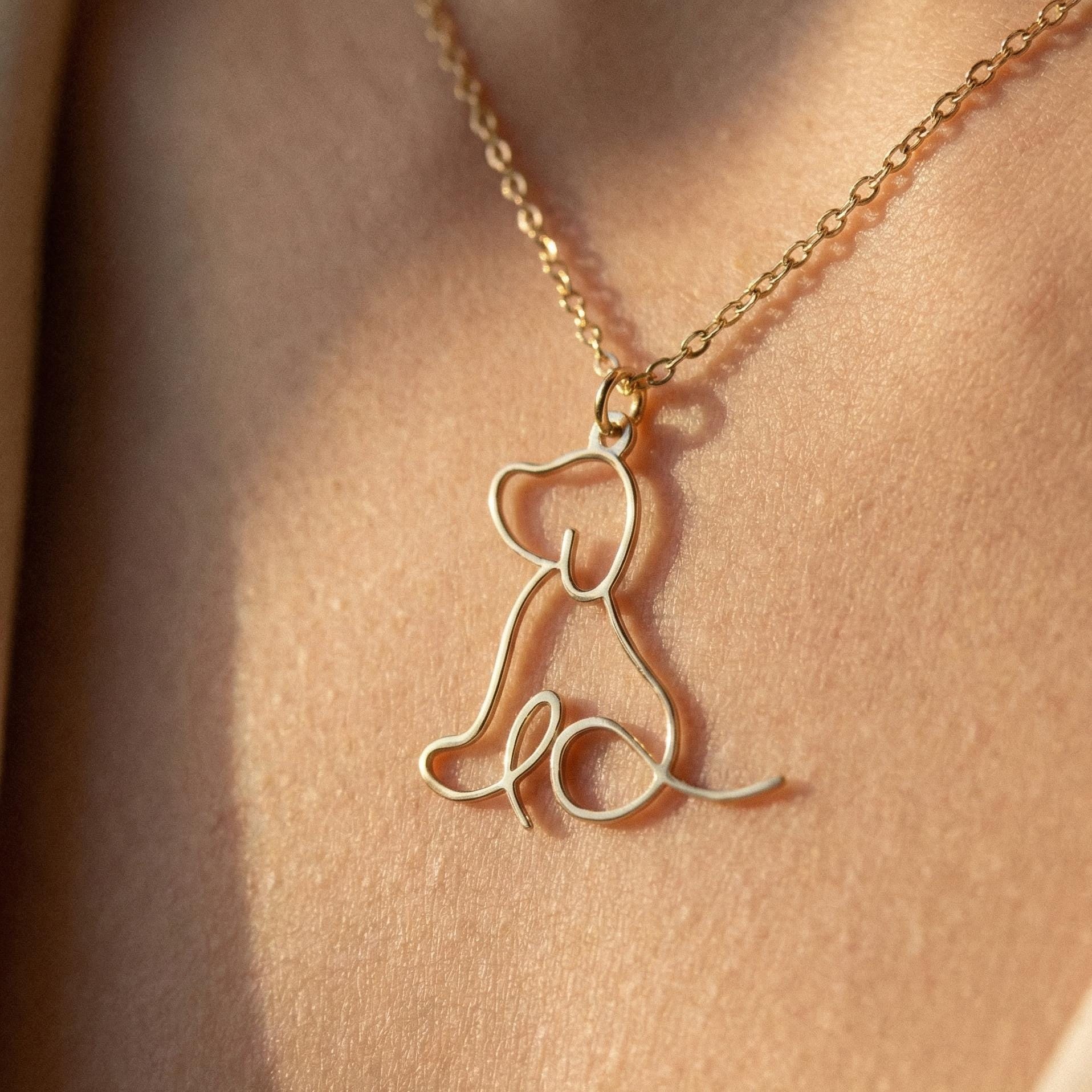 May include: A gold-colored necklace featuring a pendant in the shape of a sitting dog. The dog is depicted in a minimalist, line-art style. The necklace is worn against a skin-toned background, highlighting the delicate design.