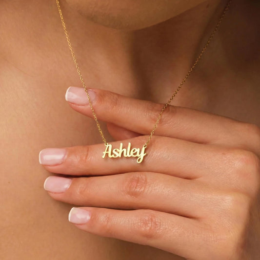 May include: A gold-colored necklace with the name "Ashley" in a cursive font. The necklace is on a delicate chain. The name is the focal point, and the necklace is displayed on a person's skin.
