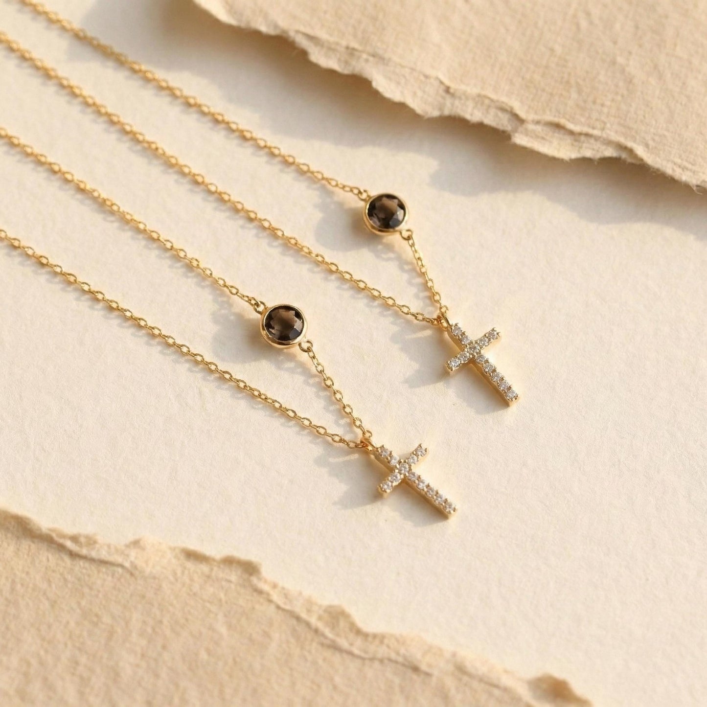Dainty gold chain necklace featuring a small pave diamond cross pendant and a round bezel-set birthstone charm, worn by a model. Personalized minimalist religious jewelry.