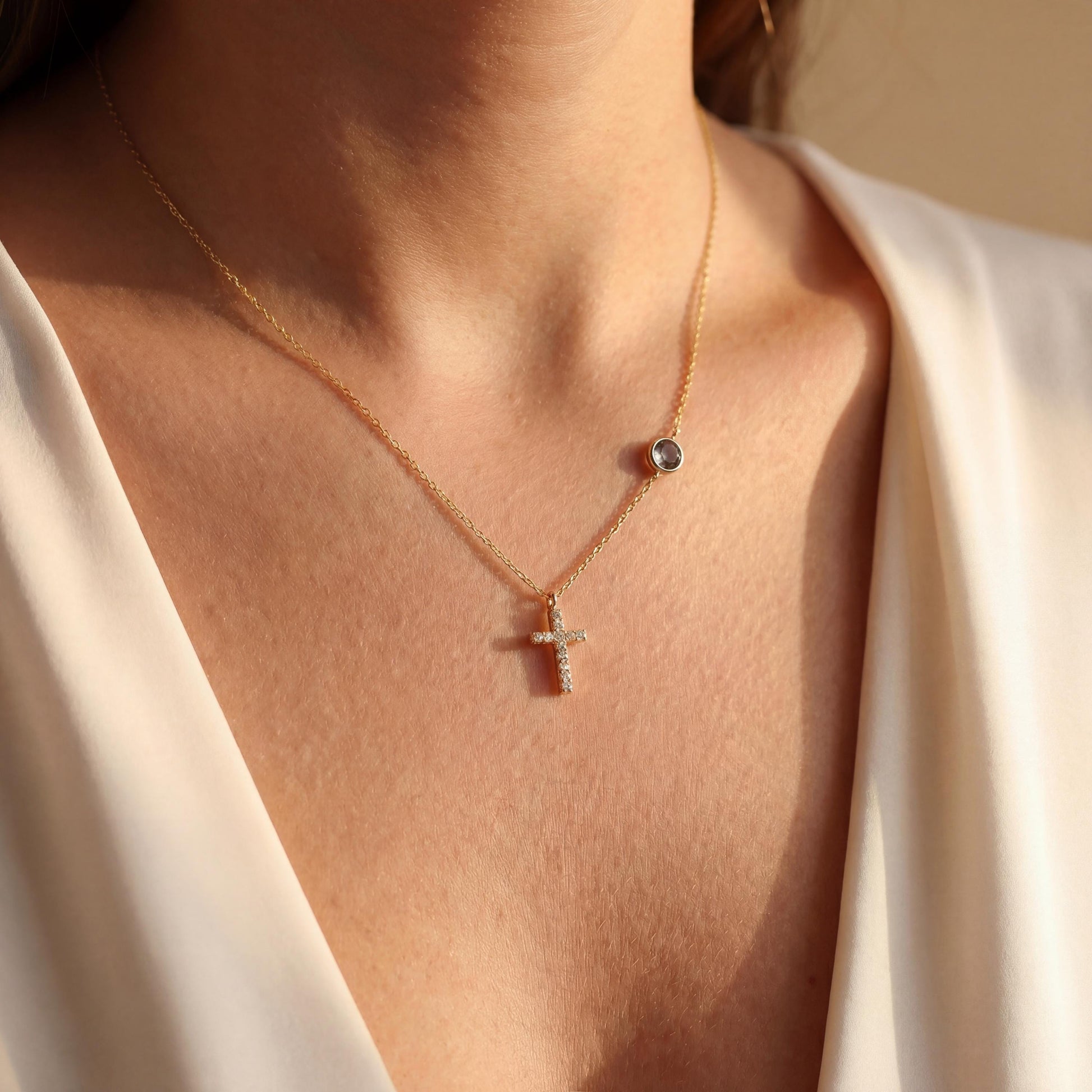 Dainty gold chain necklace featuring a small pave diamond cross pendant and a round bezel-set birthstone charm, worn by a model. Personalized minimalist religious jewelry.