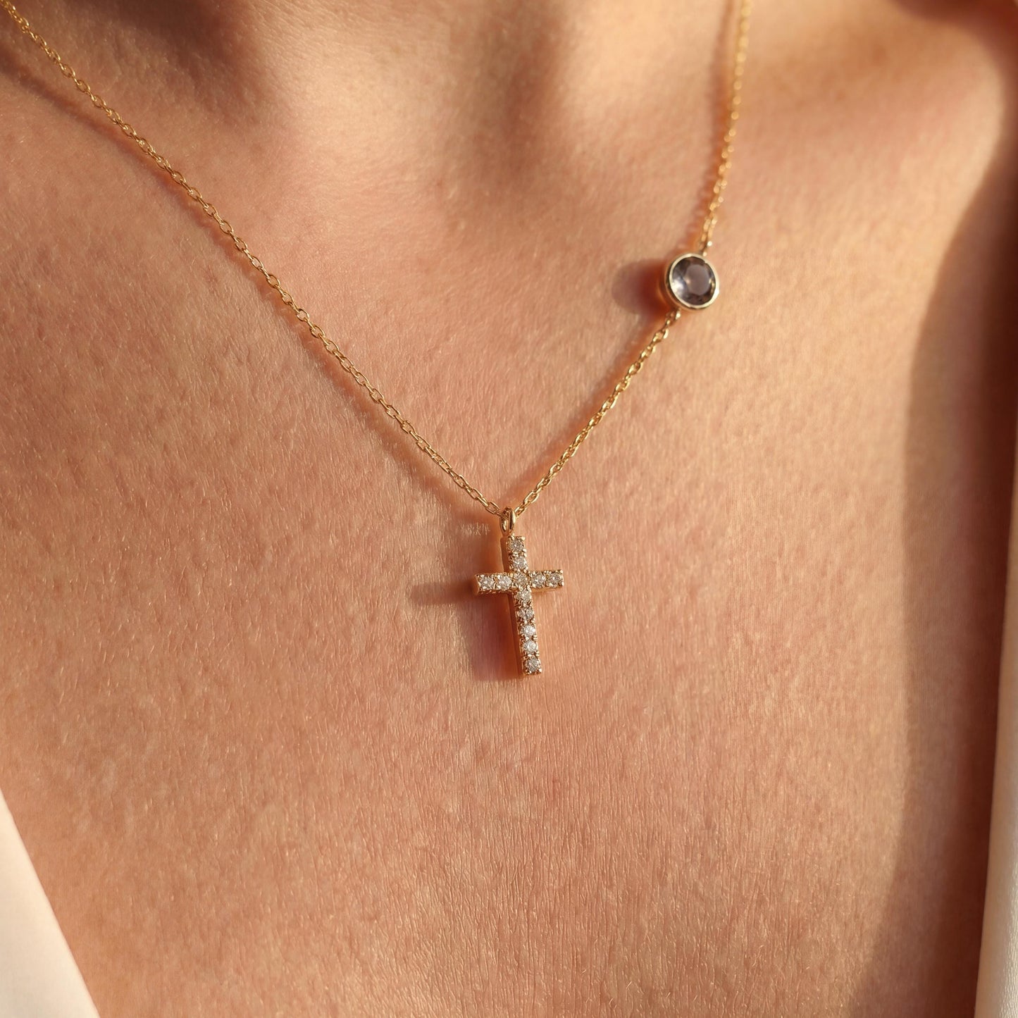 Dainty gold chain necklace featuring a small pave diamond cross pendant and a round bezel-set birthstone charm, worn by a model. Personalized minimalist religious jewelry.