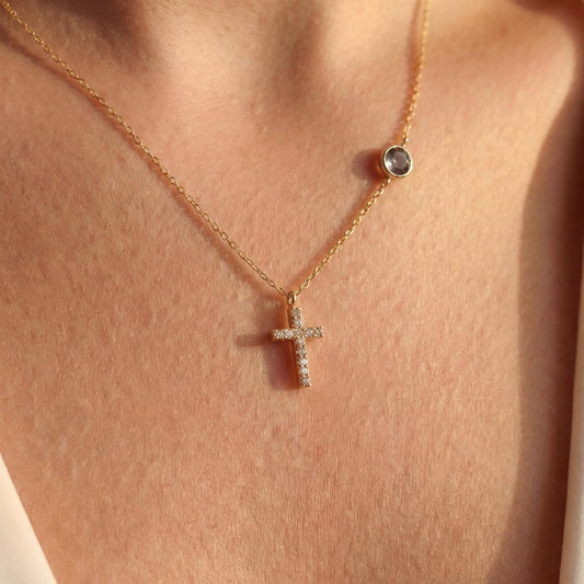 Dainty gold chain necklace featuring a small pave diamond cross pendant and a round bezel-set birthstone charm, worn by a model. Personalized minimalist religious jewelry.