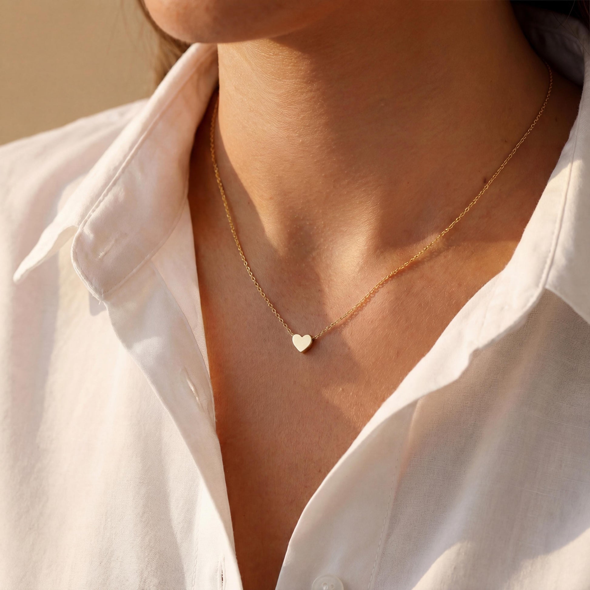 Minimalist 14k gold plated tiny puff heart necklace on a model wearing a white shirt, dainty gold choker chain detail, small heart pendant close up, perfect mothers day gift or birthday jewelry for her.