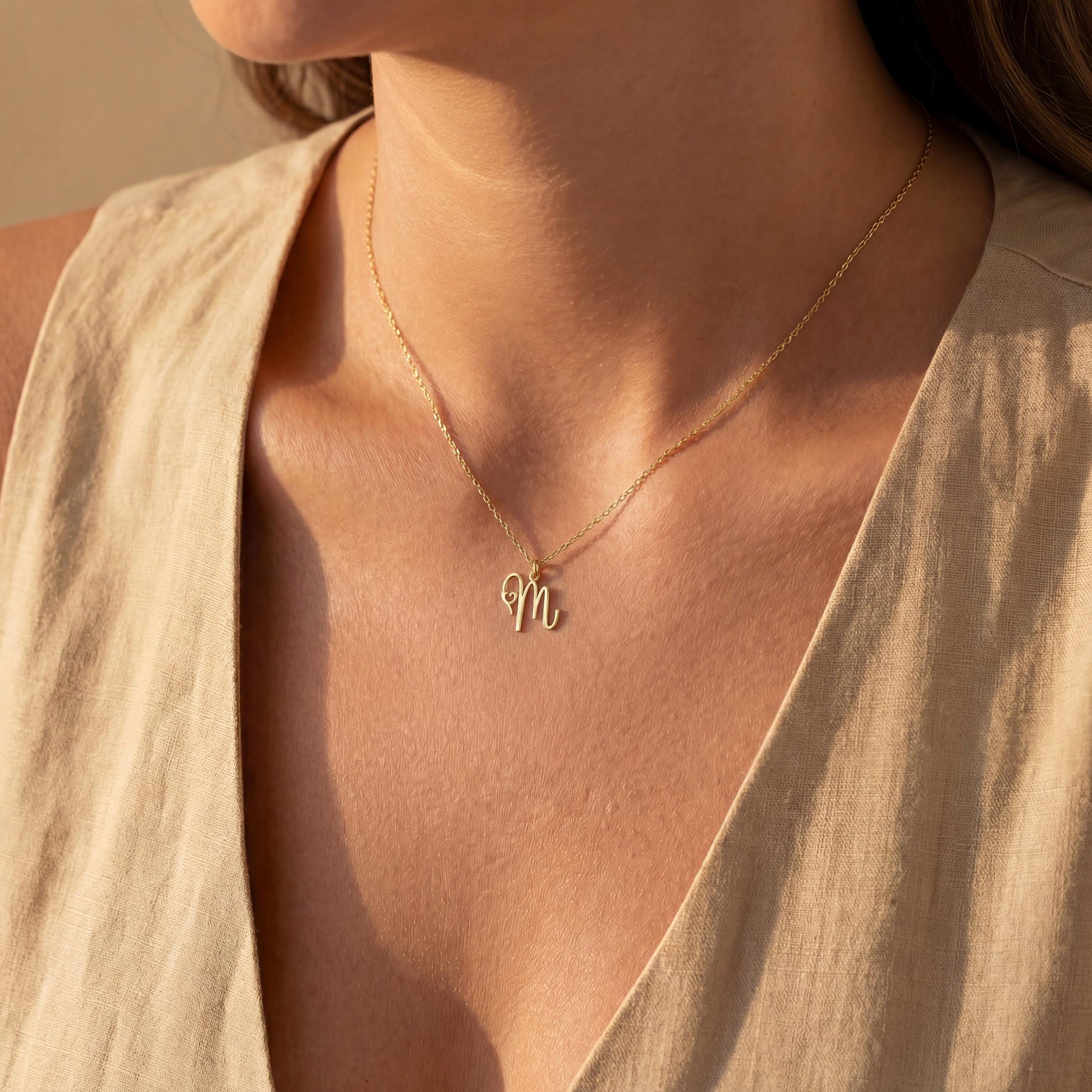 Dainty custom initial necklace with a delicate heart detail next to a cursive letter M, worn by a smiling woman in sunlight. Minimalist personalized elegant pendant.