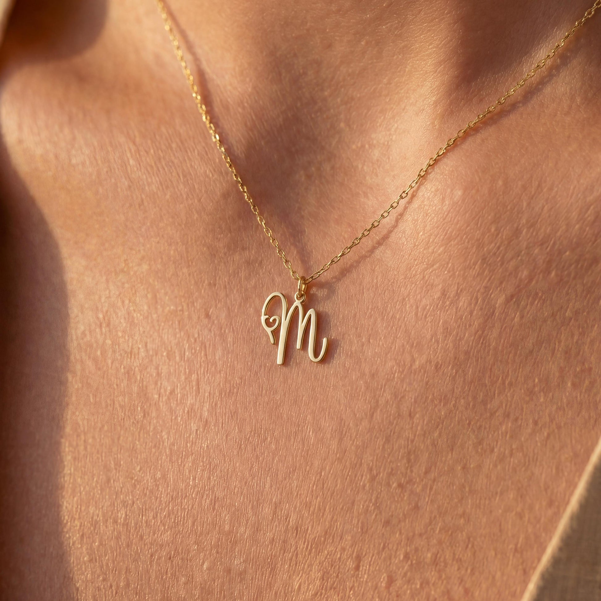 Dainty custom initial necklace with a delicate heart detail next to a cursive letter M, worn by a smiling woman in sunlight. Minimalist personalized elegant pendant.