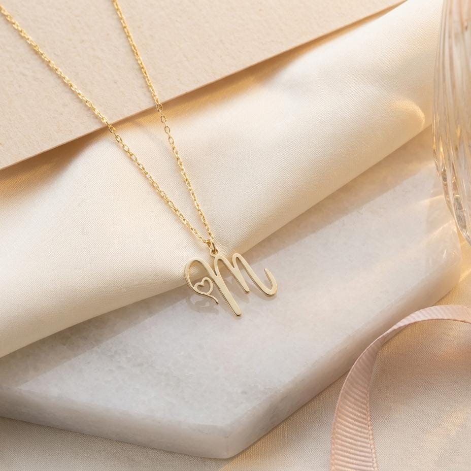 Dainty custom initial necklace with a delicate heart detail next to a cursive letter M, worn by a smiling woman in sunlight. Minimalist personalized elegant pendant.