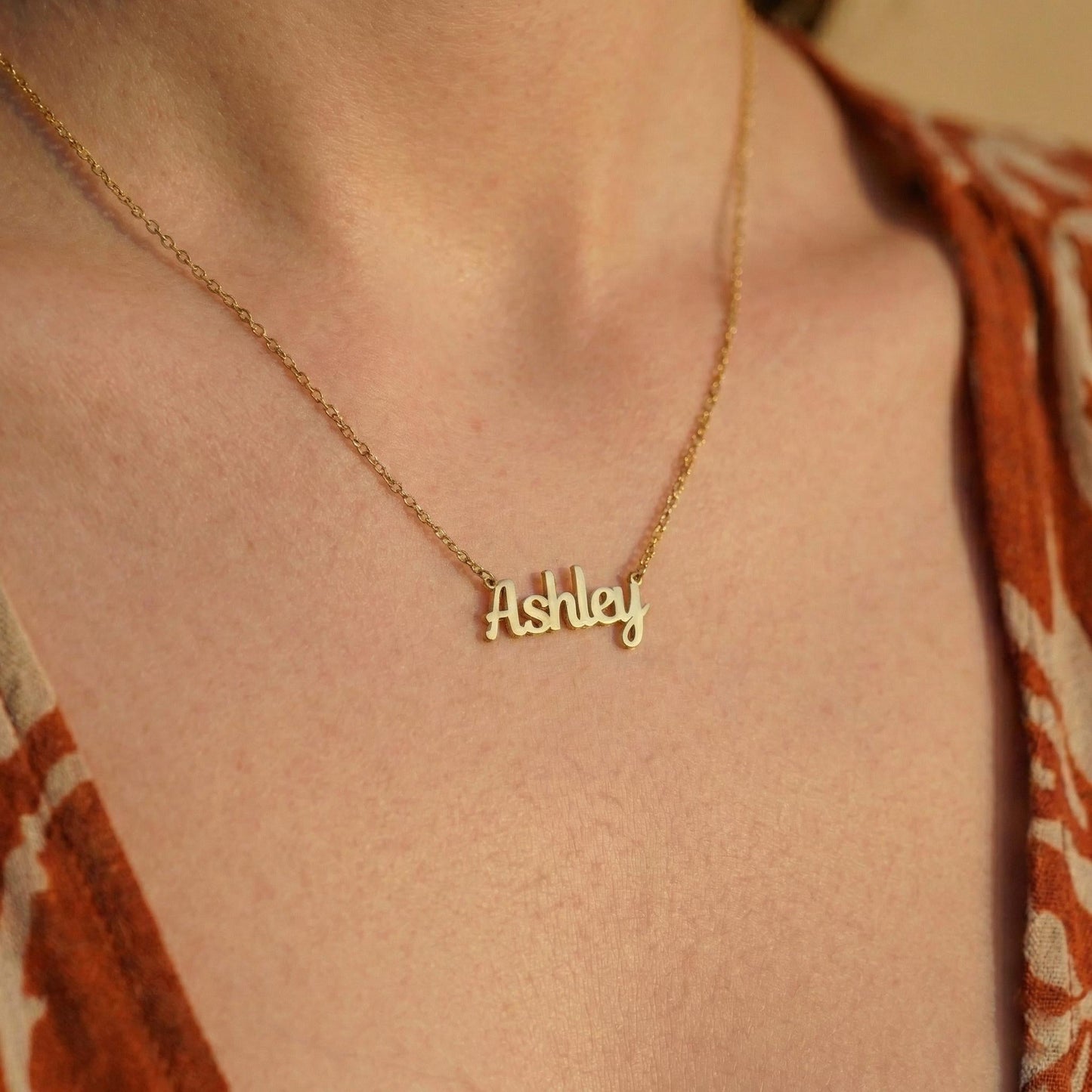 May include: A gold-colored necklace with the name "Ashley" in a cursive font. The necklace is worn around a person's neck, with the chain and pendant visible. The background is a patterned orange and brown fabric.