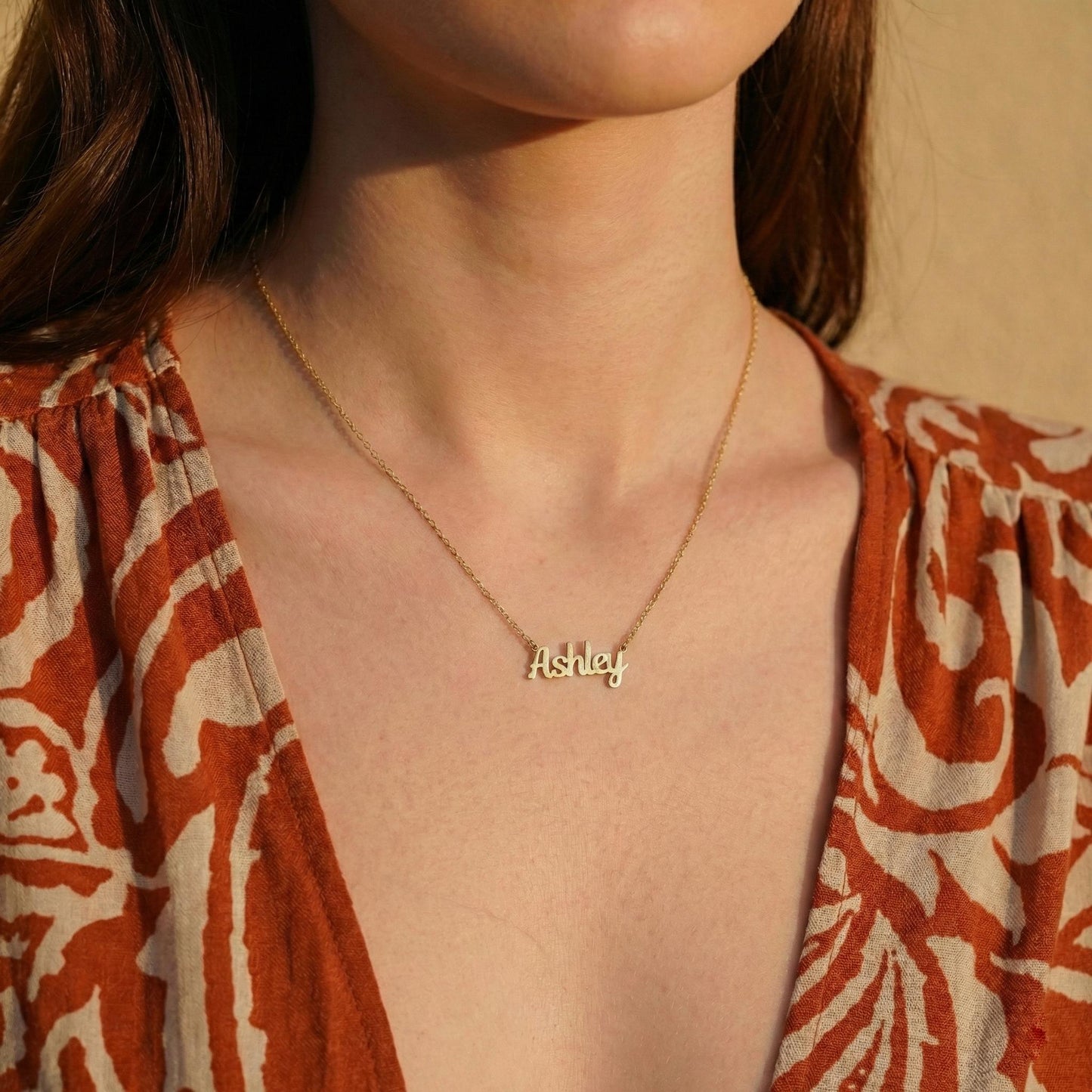 May include: A gold-colored necklace with the name "Ashley" in a cursive font. The necklace is worn against a patterned orange and white top.
