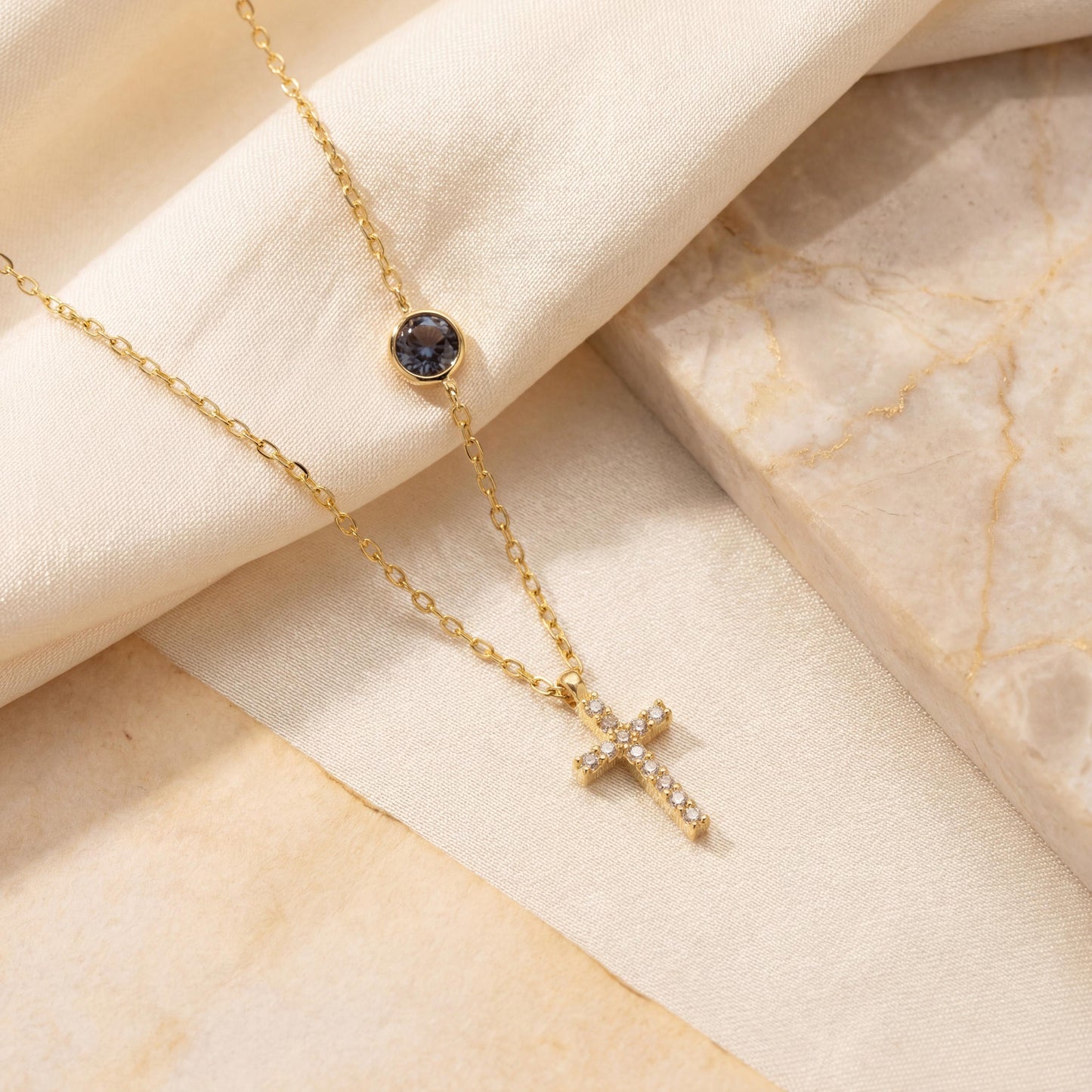 Dainty gold chain necklace featuring a small pave diamond cross pendant and a round bezel-set birthstone charm, worn by a model. Personalized minimalist religious jewelry.