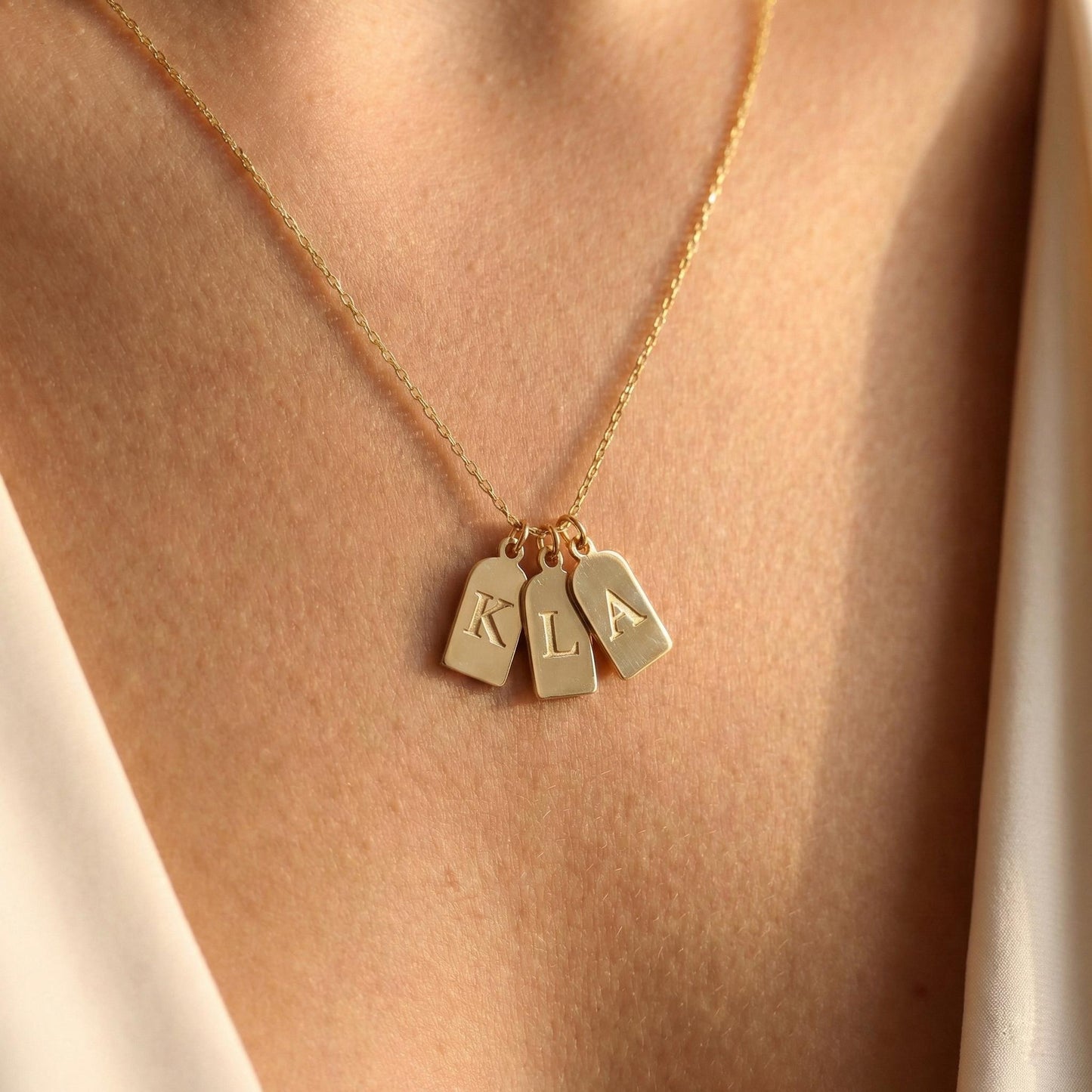 Gold initial necklace featuring three small rectangular pendant tags engraved with letters K L A, worn on a woman's neck, minimalist personalized jewelry for mom