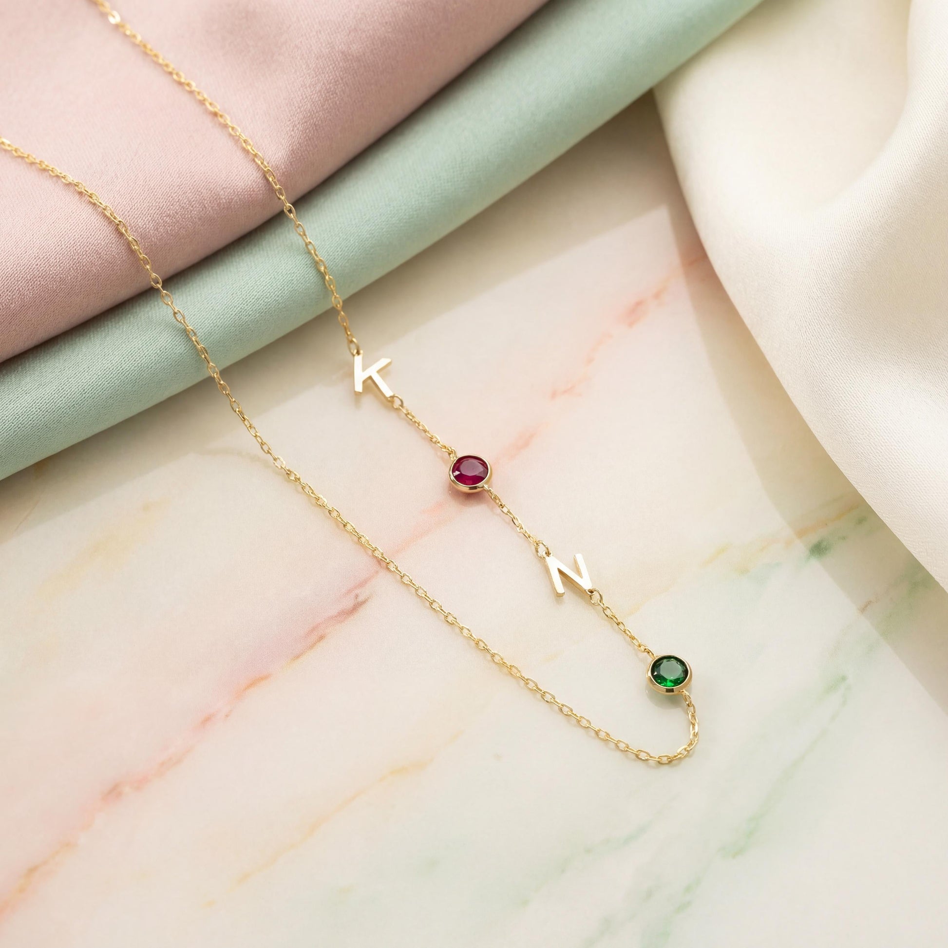 Dainty gold necklace featuring sideways initials and bezel set birthstones, personalized family jewelry for mother's day gift, minimalist custom letter necklace with ruby and emerald colored stones on neck.