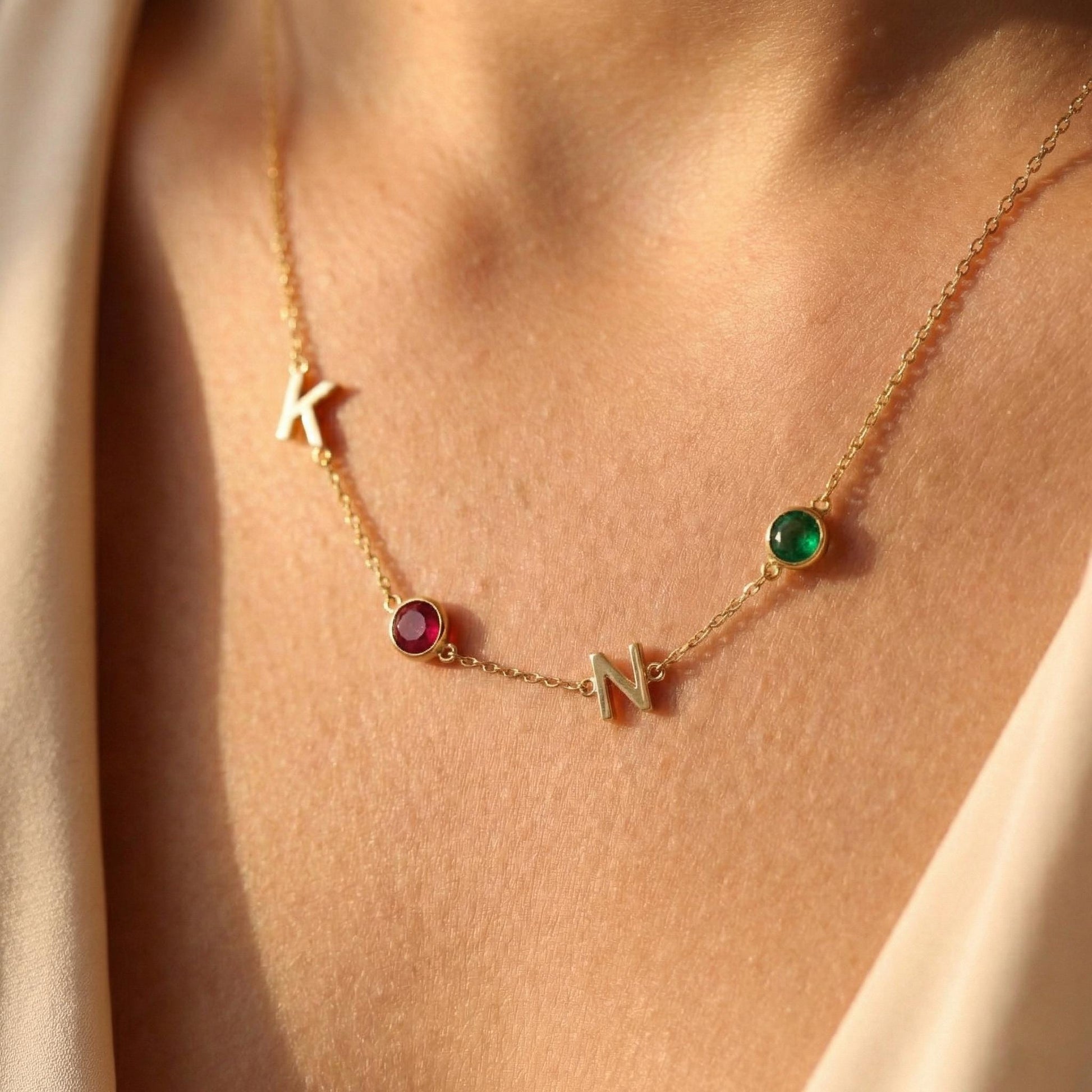 Dainty gold necklace featuring sideways initials and bezel set birthstones, personalized family jewelry for mother's day gift, minimalist custom letter necklace with ruby and emerald colored stones on neck.