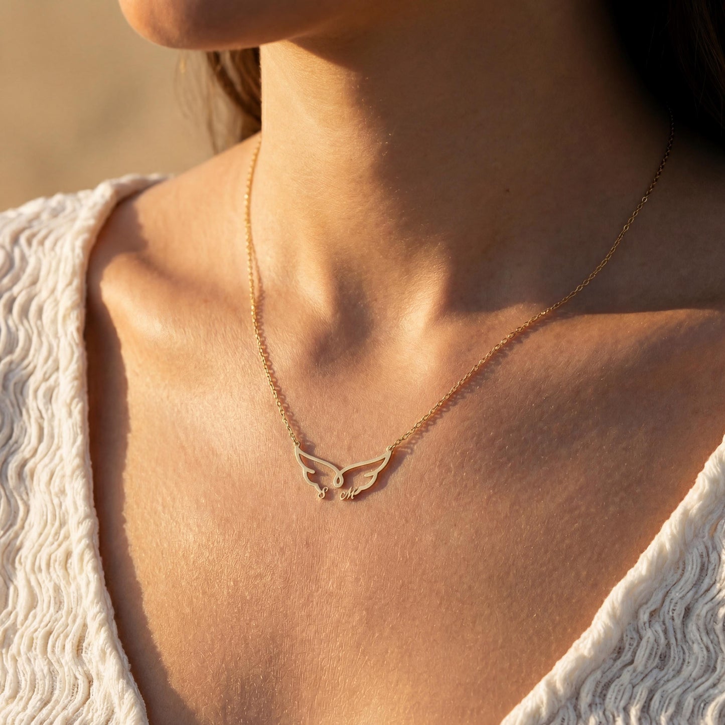 Dainty custom angel wing necklace featuring two personalized initial letters nesting at the bottom of the wings, worn on a woman's collarbone during golden hour.