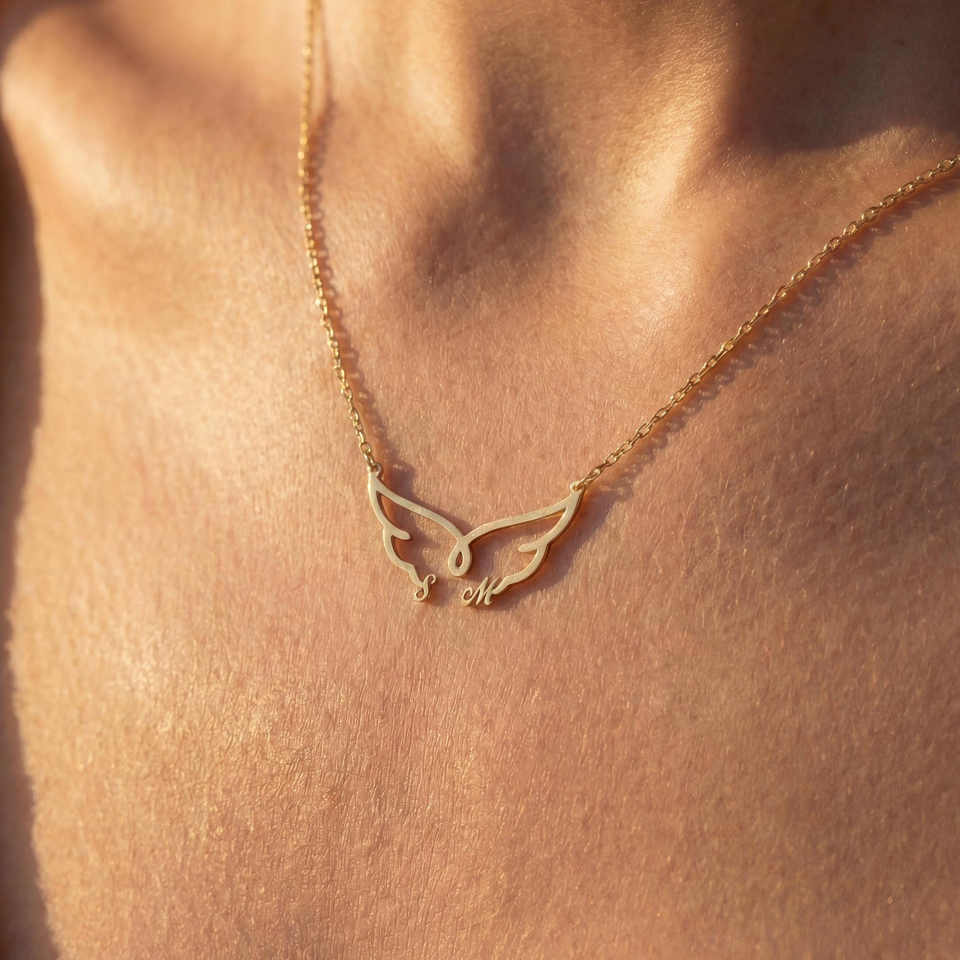 Dainty custom angel wing necklace featuring two personalized initial letters nesting at the bottom of the wings, worn on a woman's collarbone during golden hour.