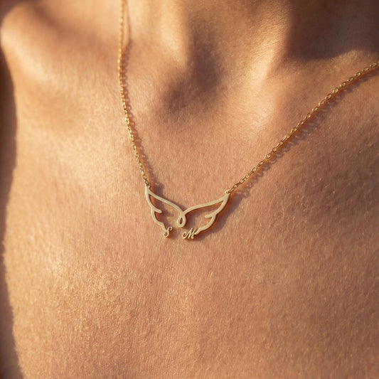 Dainty custom angel wing necklace featuring two personalized initial letters nesting at the bottom of the wings, worn on a woman's collarbone during golden hour.