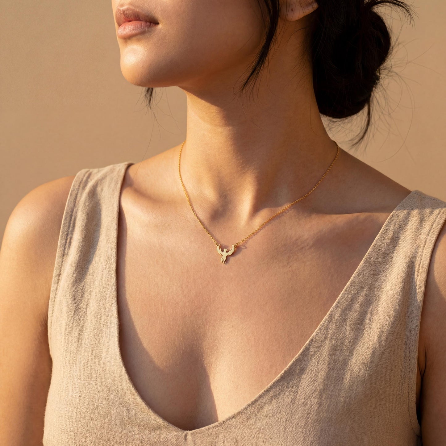 Minimalist gold phoenix pendant necklace worn on a woman's collarbone in warm sunlight, symbolizing rebirth and strength, ideal for Mothers Day gifting