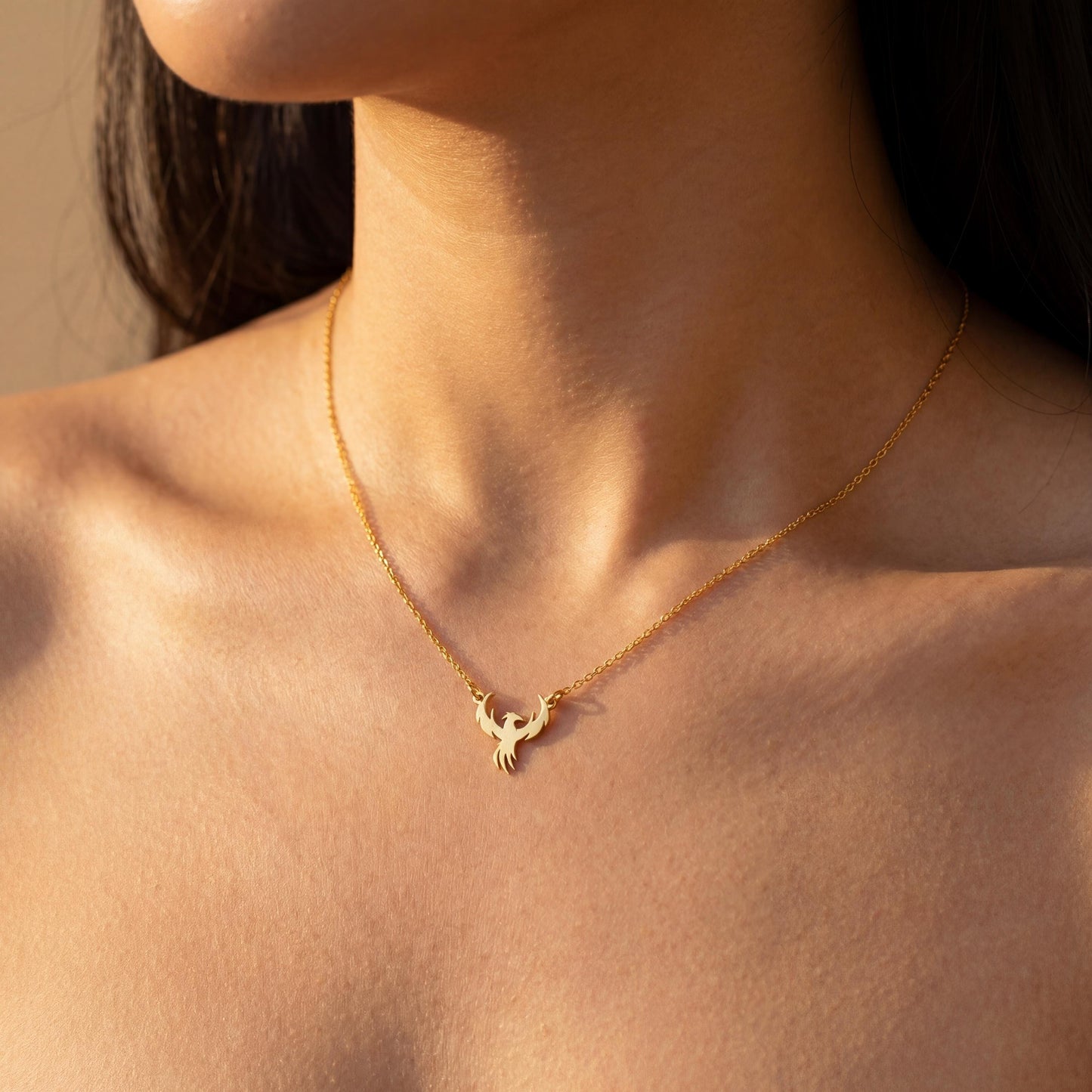 Minimalist gold phoenix pendant necklace worn on a woman's collarbone in warm sunlight, symbolizing rebirth and strength, ideal for Mothers Day gifting