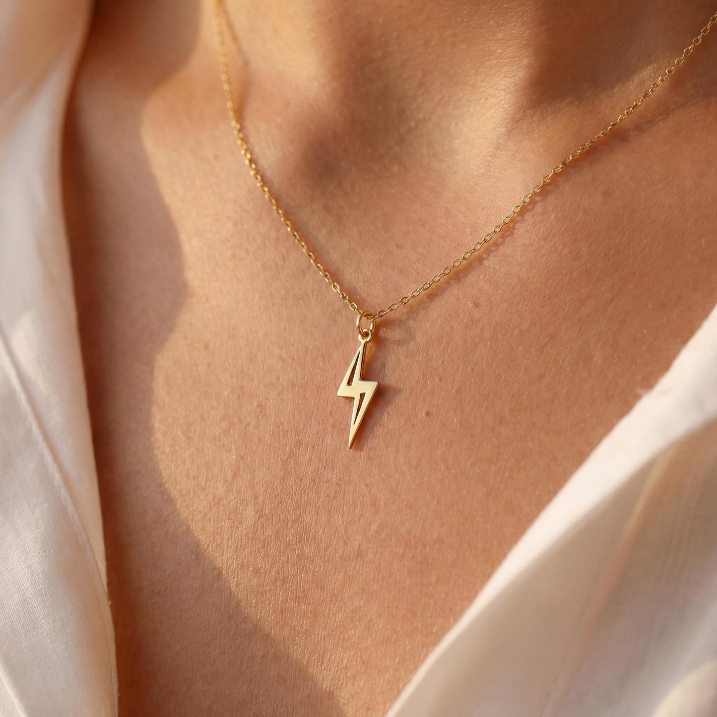 Close-up of a delicate minimalist 14k gold plated lightning bolt pendant necklace worn by a woman in a white shirt, glowing in soft sunlight.