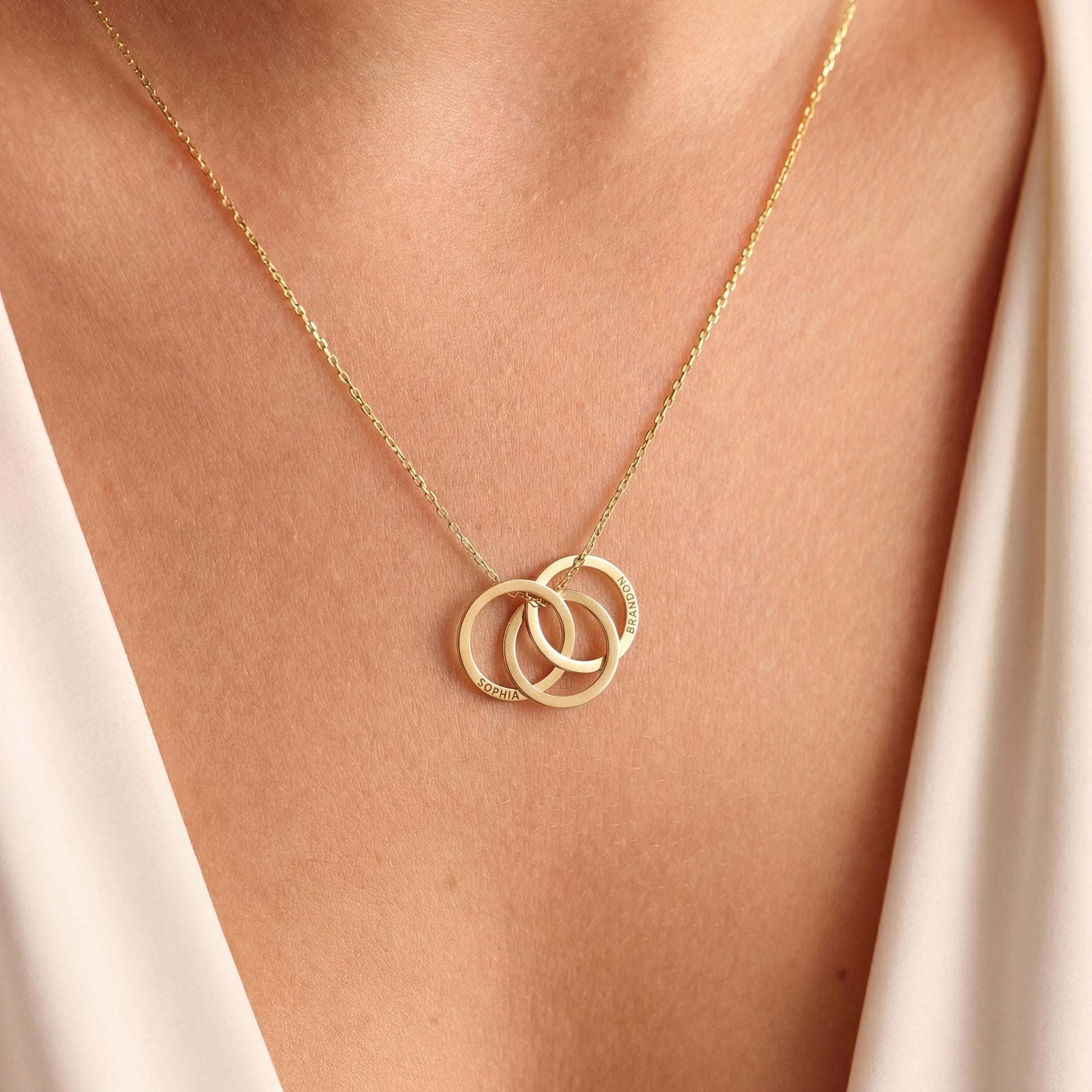 14K gold plated interlocking circle necklace featuring three beautifully intertwined rings, delicately custom engraved with the family names Sophia and Brandon, resting elegantly on a woman's collarbone.